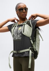 Γυναίκα με γυαλιά ηλίου ρυθμίζει τα λουριά σε ανοιχτοπράσινο σακίδιο πεζοπορίας πάνω από μαύρο γραφικό μπλουζάκι και ελαι πράσινο κολάν, μπροστά σε λευκό φόντο.