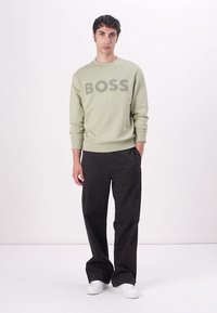 Sudadera verde claro con "BOSS" estampado en gris oscuro. Combinada con pantalones negros holgados y zapatillas blancas. Diseño simple y casual.