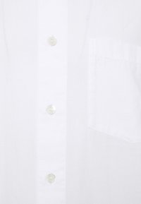 Camicia bianca con bottoni in tessuto di cotone, caratterizzata da una tasca, sei bottoni con una texture liscia e un design semplice.