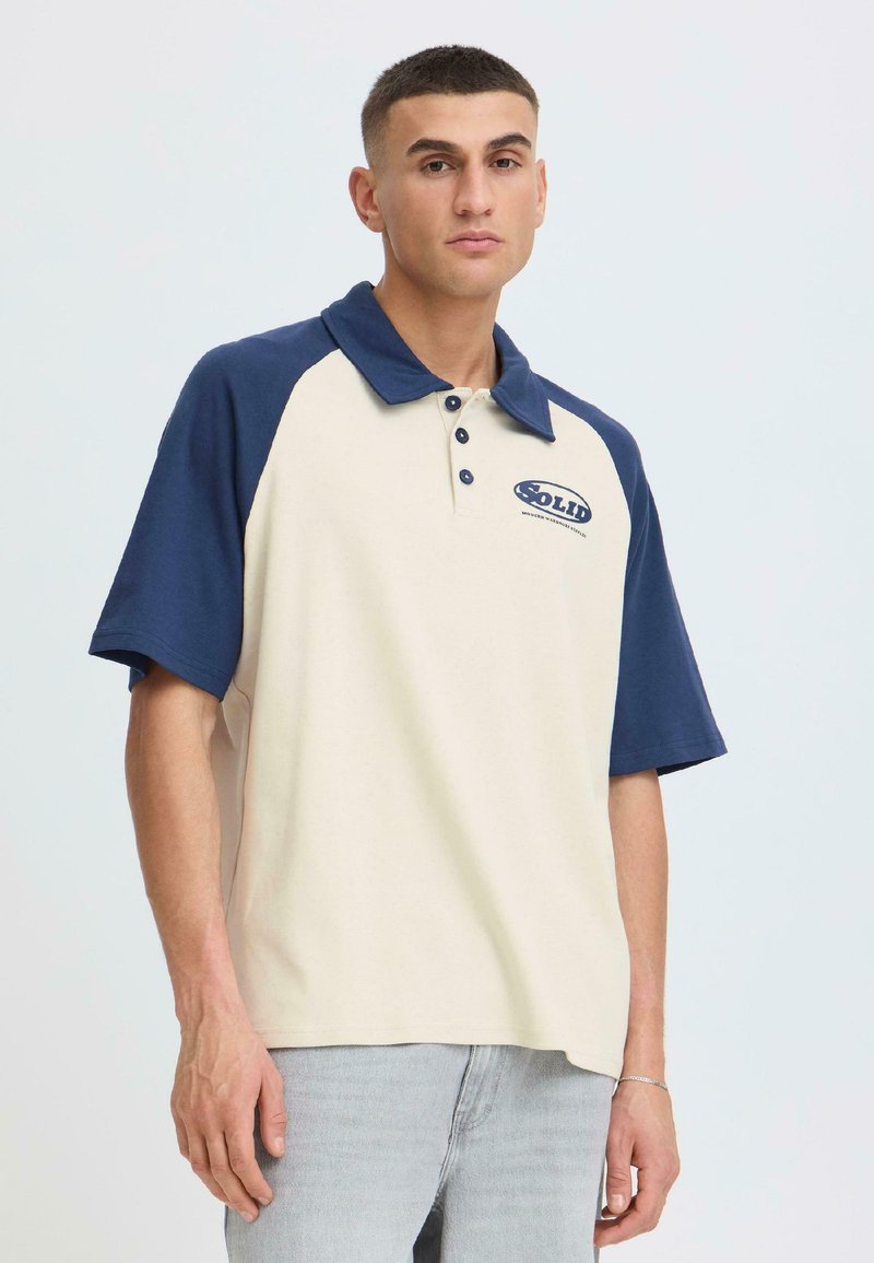 Jeune homme aux cheveux courts portant un polo à manches courtes crème et marine avec le logo "Solid" et un jean gris clair, devant un fond uni.