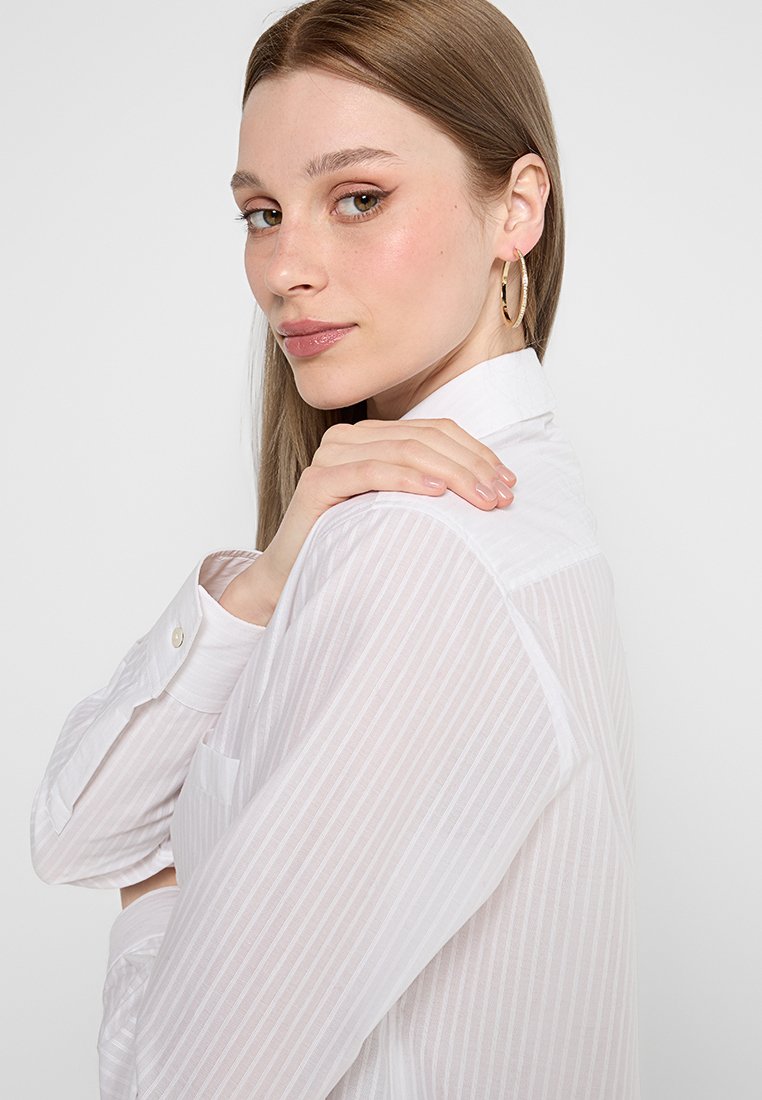 Camisa de botones blanca a rayas con mangas largas, que presenta un cuello y un bolsillo. El modelo tiene un fondo neutro, que muestra una textura suave y un ligero brillo.