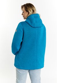 Giacca in pile blu con cappuccio, caratterizzata da una superficie testurizzata e una patch con logo sulla manica. Abbinata a pantaloni in denim chiaro.