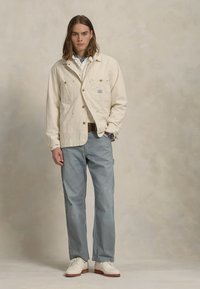Ljusbeige canvasjacka med gyllene knappar, i kombination med blå och vit randiga byxor. Vita skor fulländar outfiten.