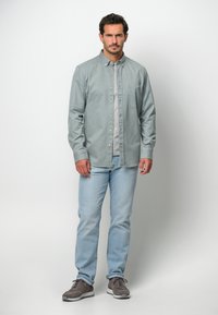 Camicia verde chiaro con bottoni e tasca, abbinata a jeans azzurri e sneakers grigie. La camicia ha maniche lunghe e colletto.