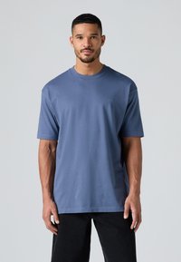 T-shirt i bomuld med rund hals i dæmpet blå farve med korte ærmer, med et diskret logo på brystet og en afslappet pasform. Sat sammen med sorte bukser.