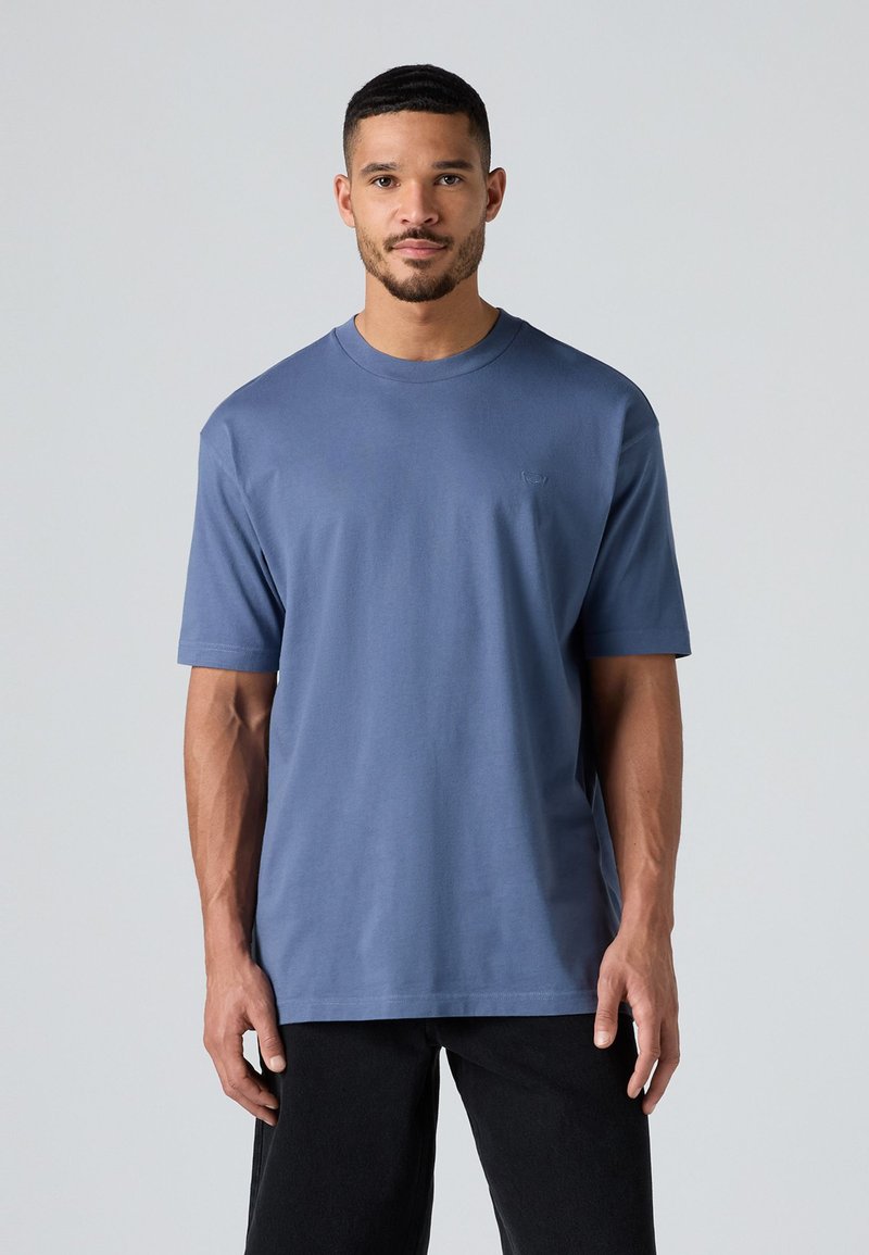 Camiseta de algodón de cuello redondo en azul apagado con mangas cortas, que presenta un logo sutil en el pecho y un corte relajado. Combinada con pantalones negros.