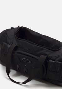 Musta duffel-laukku, joka on valmistettu kestävästä kankaasta. Laukussa on vetoketjullinen yläosa, useita taskuja ja pehmustetut olkahihnat helppoa kantamista varten.