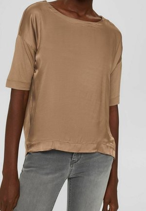 Donkerhuidige persoon draagt een losse beige satijnen top met korte mouwen en lichtgrijze aansluitende spijkerbroek tegen een effen achtergrond.