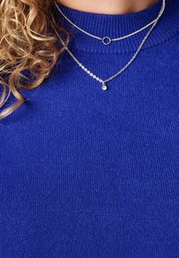 Koningblauwe gebreide trui met een hoge geribbelde kraag. Twee zilveren kettingen in laagjes; een bevat een kleine hanger.