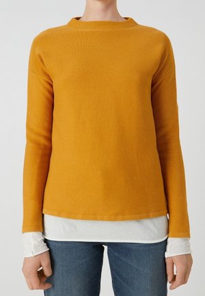 Personne portant un pull à manches longues jaune moutarde sur une chemise blanche, associé à un jean bleu, debout devant un fond gris uni.