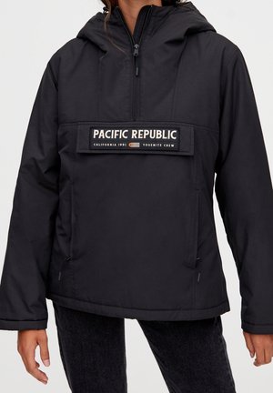 Persona que lleva una chaqueta negra con capucha, cremallera frontal y parche "Pacific Republic California 1991 Yosemite Crew" en el pecho.