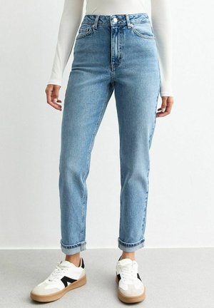 Jasnoniebieskie spodnie jeansowe z podwiniętymi nogawkami i wysokim stanem, noszone z białymi trampkami i białą długą bluzką, stojące na szarej podłodze.