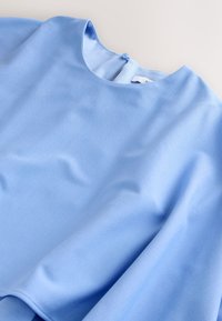 Blouse bleu clair avec encolure ronde et fermeture par boutons au dos, affichée à plat sur une surface blanche.