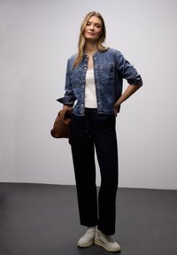 Jeune femme portant une veste en denim bleu, un haut blanc, un pantalon noir, des baskets blanches, tenant un sac marron, debout sur un sol gris devant un mur clair uni.