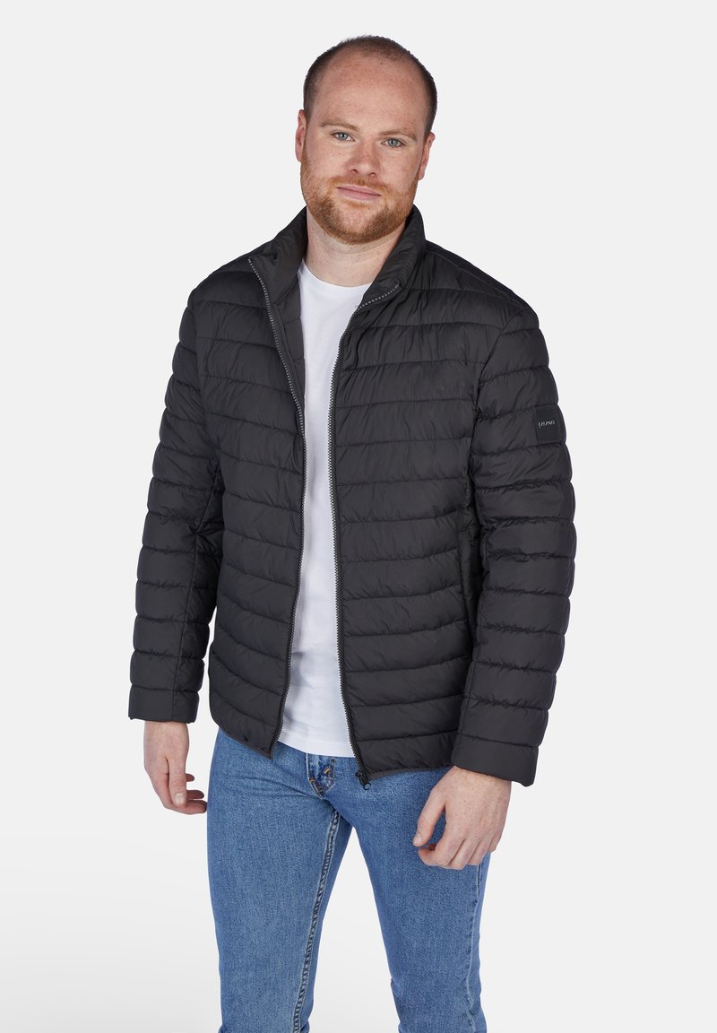 Sort pufferjakke med horisontalt quiltet design, frontlynlås og standkrave, parret med en hvid T-shirt og blå jeans.