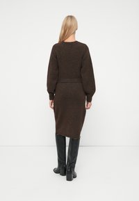 Donna con lunghi capelli biondi, che indossa un vestito maglione marrone scuro e stivali neri fino al ginocchio, in piedi davanti a uno sfondo bianco uniforme.