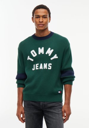 Mann trägt dunkelgrünen Pullover mit marineblauen Akzenten und dem Text "TOMMY JEANS", kombiniert mit dunkelgrauen Jeans, steht entspannt.