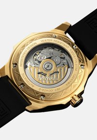 Goldfarbene Armbanduhr mit transparentem Gehäuseboden, schwarzem Gummiarmband, filigranem Muster und einer Wasserdichtigkeitsbewertung von 100 m.