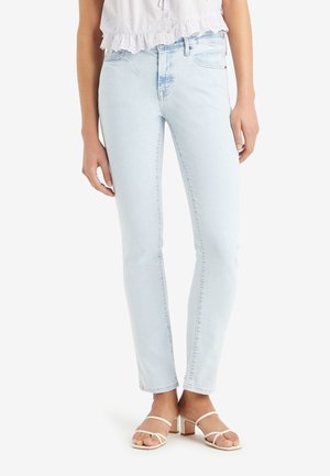 Femme portant un jean skinny bleu clair, un chemisier blanc court à œillets et des sandales blanches à talons carrés avec des brides, debout sur fond blanc.