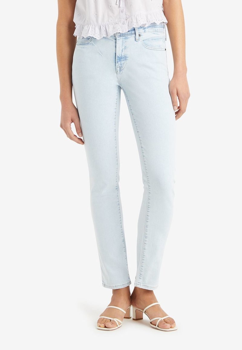 Femme portant un jean skinny bleu clair, un chemisier blanc court à œillets et des sandales blanches à talons carrés avec des brides, debout sur fond blanc.