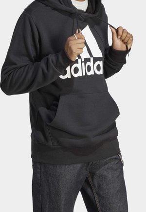 Personne portant un sweat à capuche noir Adidas avec logo blanc, tenant les cordons du sweat, associé à un jean bleu foncé sur un fond uni.
