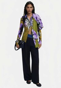 Chemise florale multicolore en vert, violet et blanc avec manches courtes, associée à un pantalon noir taille haute et un sac à main noir.