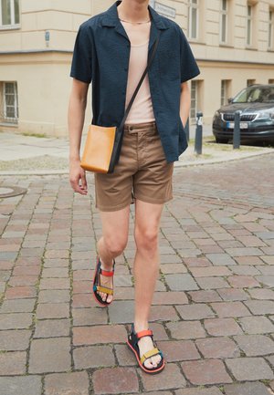Persona caminando por una calle empedrada, vistiendo una camisa de manga corta azul marino, top beige, pantalones cortos marrones, sandalias coloridas y llevando un bolso de hombro color beige.