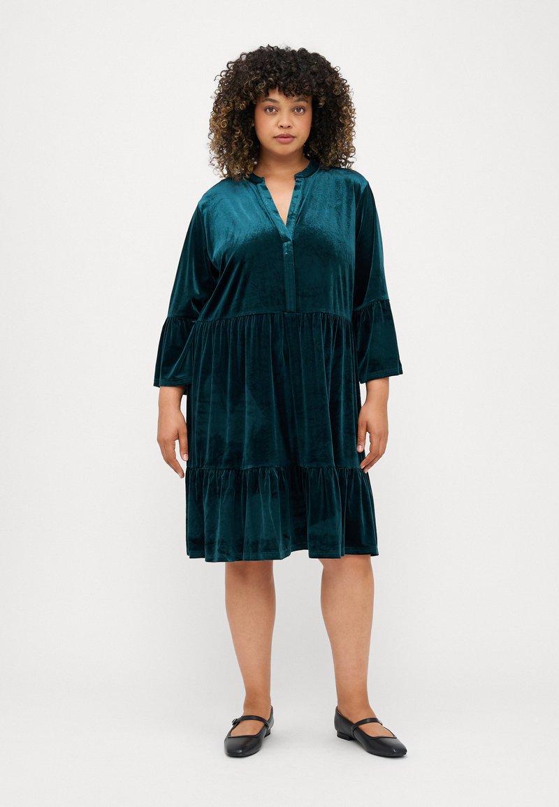 Tīrkultūru samta kleita ar V veida izgriezumu, trim ceturtdaļām garām piedurkn�ēm un pakāpenisku čokurošanas apakšmalu. Gluda tekstūra un viegls materiāls.