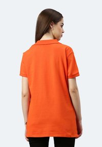 Frau mit glatten braunen Haaren, die ein oranges Kurzarm-Poloshirt trägt und mit dem Rücken zur Kamera vor einem einfarbigen hellen Hintergrund steht.