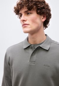 Νέος με σγουρά μαλλιά, φορώντας γκρι μακρυμάνικη μπλούζα πόλο με το λογότυπο "BOSS" στο στήθος, κοιτώντας ελαφρώς προς τα αριστερά.