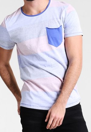 Homme portant un t-shirt à manches courtes avec des rayures pastel horizontales et une poche poitrine bleue, associé à un pantalon noir.