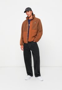 Brown fleece jacket with a contrasting brown nylon front, featuring a zip closure and a high collar, paired with black jeans and white sneakers.