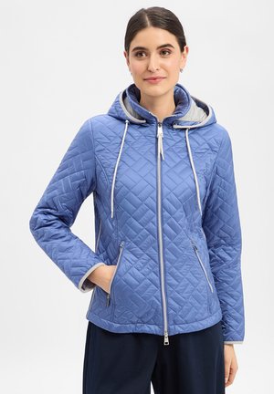 Vrouw draagt een lichtblauw doorgestikt jack met ritssluiting, capuchon en trekkoorden, handen in de zakken, tegen een effen lichte achtergrond.