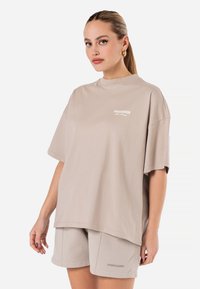 Bežové oversized tričko s krátkymi rukávmi a okrúhlym výstrihom; s bielym logom na hrudi. Vytvorené v páre s vyhovujúcimi bežovými šortkami.