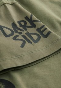 Olivově zelené bavlněné tričko s velkým černým grafickým nápisem "DARK SIDE". Tkanina má hladkou texturu a viditelné detaily šití.