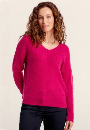 Pull en V en peluche rose avec des manches longues, à la coupe décontractée et à l'ourlet côtelé. Porté sur un jean foncé. Texture douce visible.