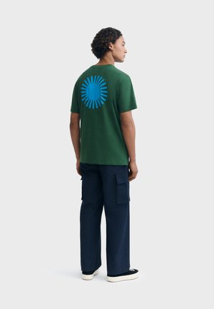 Homme portant un t-shirt vert foncé avec un motif d'éclat de soleil bleu dans le dos, un pantalon cargo bleu marine et des baskets noires avec des semelles blanches, de profil.