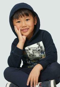 Ensemble hoodie et pantalon de survêtement bleu marine avec un imprimé graphique sur la poitrine, texture douce et poignets côtelés. Pose assise sur un fond clair.