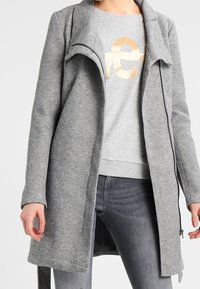 Abrigo gris con cremallera frontal, cuello abierto, tejido texturizado, bolsillos laterales y mangas largas, combinado con una sudadera gráfica gris claro.