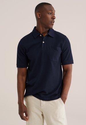 Homme portant un polo bleu marine à manches courtes avec poche, pantalon beige, regardant vers la droite, mains détendues le long du corps sur fond uni.