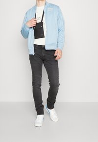 Giacca azzurro chiaro con zip e dettagli bianchi, borsa nera a tracolla, t-shirt bianca con stampa grafica, jeans neri e sneakers bianche con dettagli blu.