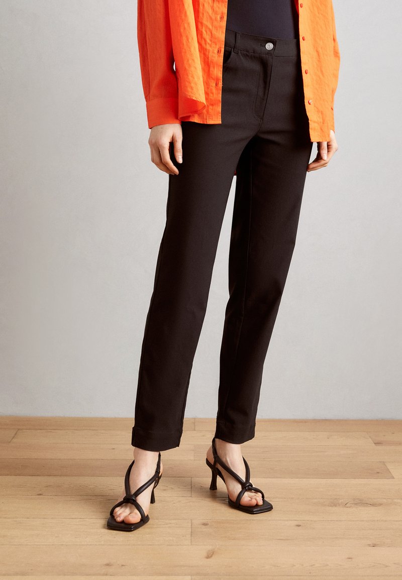 Black tailored pants with a slim fit, featuring a front button closure and a smooth texture. Paired with strappy black heels.