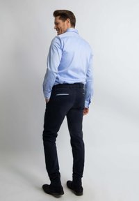Lichtblauwe overhemd met lange mouwen en knoopmanchetten, gecombineerd met donkerblauwe slim-fit broek en zwarte schoenen, gezien vanuit de achterkant. Effen achtergrond.