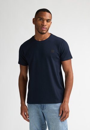 Camiseta de manga corta azul marino con textura acanalada, cuello redondo y pequeño parche con logo en el pecho; combinada con jeans de mezclilla clara.