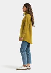 Pull polaire jaune moutarde avec un col montant et des manches longues, associé à un jean bleu clair et des baskets blanches. La texture semble douce.