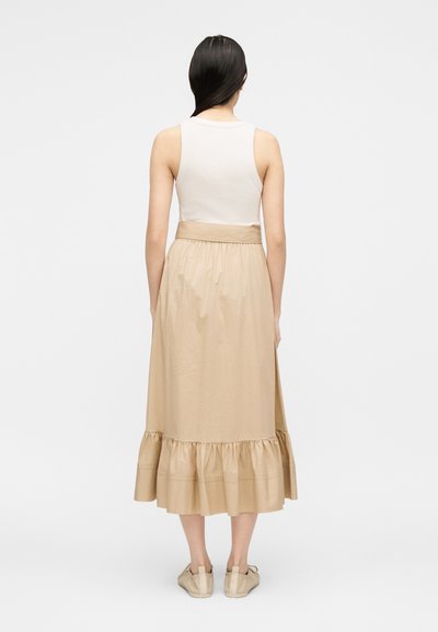 Γυναίκα με μακριά σκούρα μαλλιά, φοράει λευκό αμάνικο τοπ, μπεζ μίντι φούστα με βολάν στο τελείωμα και μπεζ παπούτσια, στέκεται γυρισμένη πλάτη.