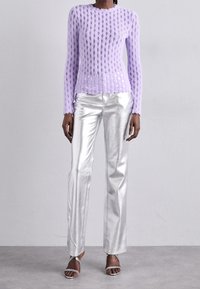 En svart person som bär en lila långärmad nättröja med silverfärgade metallbyxor och silvriga klackskor med remmar, stående mot en enfärgad vägg.