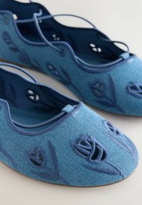 Zapatillas de ballet de tela azul con recortes florales bordados y bordes festoneados mostradas sobre un fondo claro.