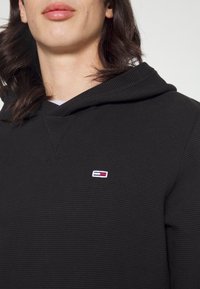 Sudadera con capucha negra hecha de tejido texturizado, con un pequeño parche de logo multicolor en el pecho.