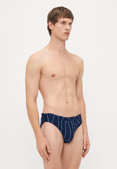 Junger Mann mit kurzen braunen Haaren, der marineblaue Badeanzüge mit einem hellblauen abstrakten Liniendesign trägt und vor einem weißen Hintergrund steht.
